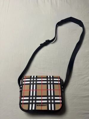 Burberry axelväska med rutigt mönster - Snygg axelväska från Burberry med klassiskt beige, svart, vitt och rött rutmönster. Väskan har justerbar svart axelrem och lock med Burberry-logga framtill. Perfekt för dig som gillar stilrena och ikoniska accessoarer.