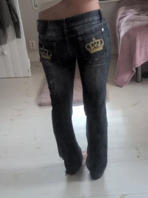 Mörkblå Viktoria Beckham jeans med guldkronor - Säljer ett par mörkblå lite boutcut men strait jeans med snygga guldfärgade kronor på bakfickorna. Jeansen har låg midja. Perfekta för dig som vill sticka ut lite extra med detaljer på baksidan. Benlängden är runt 77cm och runt midjan 30cm. Skulle säga att byxorna är i storlek Medium. 