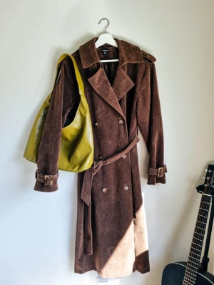 Brun vintage trenchcoat i mockaimitation S 90tal 90s grunge - Snygg brun trenchcoat i mockaimitation med dubbelknäppning, bred krage och skärp i midjan. Jackan har axelklaffar och justerbara spännen vid ärmsluten för extra stilpoäng. Använd en gång, perfekt skick 