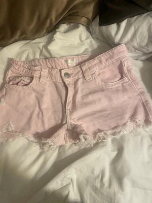 Ljusrosa  shorts  - Säljer ett par ljusrosa shorts i bomull med fransiga kanter och klassisk femficksdesign. Shortsen har bälteshällor, knapp och dragkedja framtill samt fickor med lock bak. Perfekta för varma dagar och en avslappnad look.