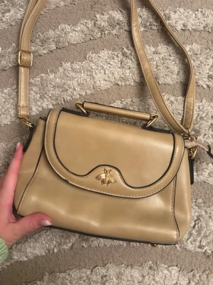 Beige handväska med gulddetaljer - Stilren beige handväska i konstläder med guldfärgad bi-detalj på locket. Väskan har både axelrem och handtag, samt magnetlås och flera fack inuti för smart förvaring. Perfekt accessoar för dig som gillar klassisk och trendig design.