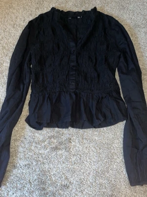 Svart blus med volang och knappar - Snygg svart blus med långa ärmar, knappar framtill och smockad överdel. Blusen har volang nertill och lätt rynkad struktur som ger en trendig look. Perfekt att styla med jeans eller kjol för en cool outfit.