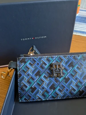 Blå mönstrad plånbok Tommy Hilfiger - Snygg plånbok från Tommy Hilfiger i blått och svart med geometriskt TH-mönster och metallogo framtill. Plånboken har dragkedja, guldfärgade detaljer och en praktisk krok för att fästa i väska eller nyckelring. Materialet är tåligt konstläder.
