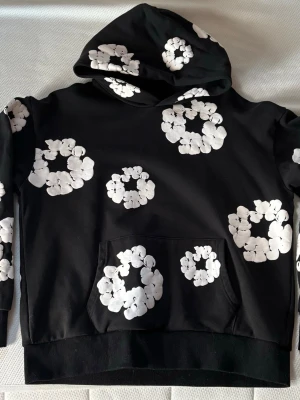 Svart Denim Tears Hoodie - Hoddien passar mer som än S än M enligt mig. Snygg svart hoodie med stora vita blommönster över hela plagget. Perfekt för dig som gillar streetwear och vill sticka ut lite extra.