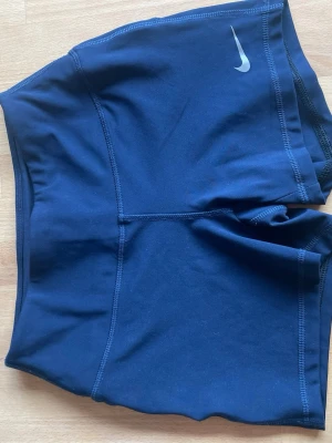 Mörkblå träningsshorts från Nike - Snygga mörkblå träningsshorts från Nike med hög midja och korta ben. Tillverkade i stretchigt funktionsmaterial som passar perfekt för gym, löpning eller annan träning. Diskret Nike-logga framtill. Sköna och flexibla för alla typer av sport.
