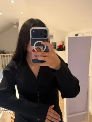 Svart långärmad blus med v-ringning - Snygg svart blus med lång ärm och v-ringad hals. Blusen har en figurnära passform och är tillverkad i ett slätt tyg som ger en stilren look. Perfekt att matcha med jeans eller kjol för en trendig outfit. En av knapparna sitter löst, går enkelt att fixa med tråd och nål! 