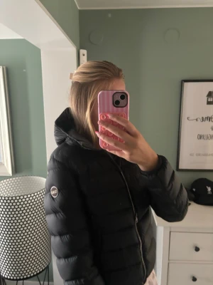 Colmar jacka  - Snygg svart dunjacka med huva och dragkedja framtill. Jackan har en puffig design och ett runt märke på ärmen. Perfekt för kalla vinterdagar och ger en stilren look.