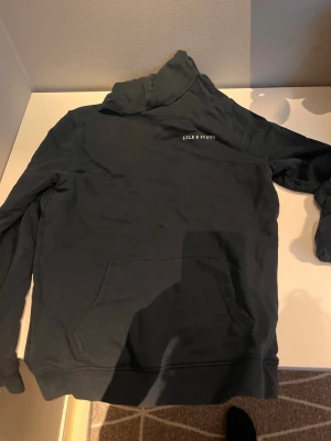 Mörkblå hoodie från Lyle & Scott - Snygg mörkblå hoodie från Lyle & Scott med klassisk huva och magficka. Diskret logga på bröstet och mjukt material som känns skönt mot huden. Perfekt för en avslappnad och cool stil.
