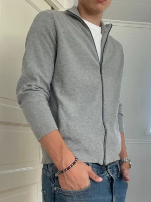 Ljusgrå ziptröja Massimo Dutti - Säljer en stilren ljusgrå ziptröja med hel dragkedja framtill. Tröjan har en smal passform och är tillverkad i ett mjukt material som passar perfekt till jeans. Långärmad och enkel design som funkar till många olika looks.