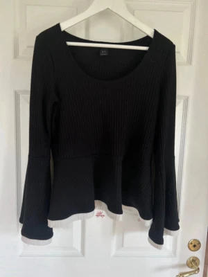 Svart ribbad topp från Lindex - Svart ribbad topp från Lindex med lång ärm och lätt utsvängd nederdel. Toppen har rund halsringning och vita detaljer längst ner på ärmar och fåll. Perfekt för dig som gillar enkel men snygg stil.