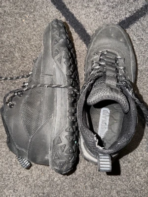 Svarta kängor från Timberland - Säljer ett par svarta kängor från Timberland med robust sula och grovt mönster för extra grepp. Skorna har snörning med metallhål och detaljer i textil och syntet. Perfekta för utomhusaktiviteter och streetwear.