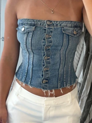 Jeanstopp Zara - Säljer en snygg blå tubtopp i denim med knappar framtill och rå kant nertill. Toppen har dekorativa fickor och sömmar som ger en korsettliknande look. Perfekt till sommaren för en trendig och cool stil.