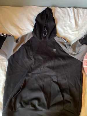 Svart och grå hoodie från Champion - Snygg svart och grå hoodie från Champion med huva och magficka. Ärmarna har gråa partier och coola band med Champion-loggor längs axlarna. Perfekt för en avslappnad streetwear-look. Materialet känns mjukt och bekvämt.