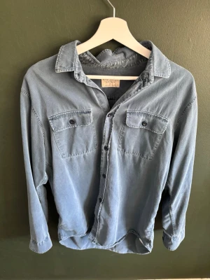 Nudie skjorta - Snygg blå skjorta med två bröstfickor och knappar framtill. Skjortan har en avslappnad, oversized passform och är gjord i mjuk bomull. Perfekt att bära öppen över en t-shirt eller knäppt för en clean look.
