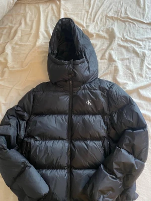 Svart pufferjacka från Calvin Klein - Snygg svart pufferjacka från Calvin Klein med stor huva och dragkedja framtill. Jackan har det klassiska CK-logotypen på bröstet och är tillverkad i ett glansigt, vadderat material som håller dig varm under kalla dagar. Perfekt för vinter och streetwear-stil. Jackan är helt ny utan prislapp!