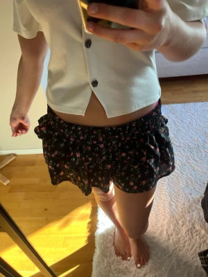 Svarta blommiga volangshorts - Säljer en svart kjol med små rosa och röda blommor. Kjolen har volangdetaljer och resår i midjan för skön passform. Perfekta för varma dagar och ger en söt och trendig look.