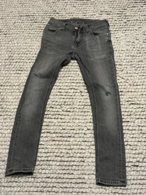 Grå slim fit jeans från Nudie Jeans - Säljer ett par grå slim fit jeans från Nudie Jeans med klassisk femficksmodell och diskreta slitningar på benen. Jeansen har en snygg tvättad look och detaljerade bakfickor med Nudie-signatur. Perfekta till en avslappnad streetstil.