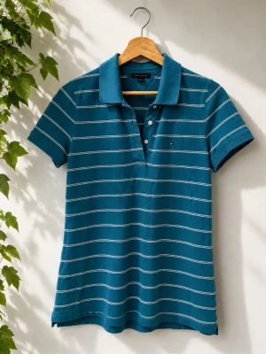Blå randig pikétröja Tommy Hilfiger (dam) - Snygg blå pikétröja från Tommy Hilfiger med vita horisontella ränder och klassisk krage. Tröjan har korta ärmar, knappar framtill och den ikoniska loggan broderad på bröstet. Perfekt för en avslappnad men stilren look.