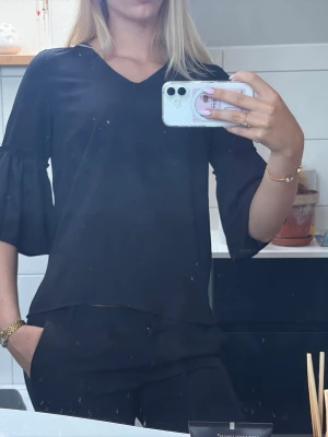 Svart topp med volangärm och v-ringning - Stilren svart topp med v-ringad hals och vida volangärmar i trekvartslängd. Tunnare material som faller snyggt och ger en avslappnad men chic look. Perfekt att matcha med både jeans och kjol för en trendig outfit.