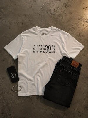 Vit Maison Margiela t-shirt med tryck - Säljer en vit t-shirt från Maison Margiela med det ikoniska siffertrycket och en markerad 6:a på bröstet. T-shirten har rund hals, korta ärmar och är gjord i mjuk bomull. Perfekt för dig som gillar minimalistisk och stilren design.