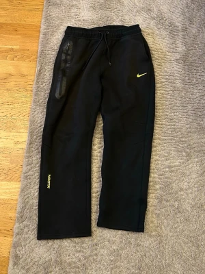 Svarta NOCTA x Nike mjukisbyxor - Snygga svarta mjukisbyxor från NOCTA x Nike med gul logga på benet och klassisk Nike Swoosh. Byxorna har dragsko i midjan, raka ben och en dragkedjeförsedd ficka. Perfekta för en chill och sportig look.