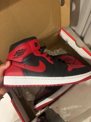 Svart och röd Air Jordan 1 Mid - Säljer ett par Air Jordan 1 Mid sneakers i svart och rött läder med klassisk logga på sidan. Skorna har svart snörning, vit mellansula och röd yttersula. Perfekta för dig som gillar streetwear och vill sticka ut med en ikonisk modell.