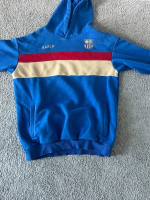 Blå Barça hoodie med röd och beige rand - En Barcelona tröja från 2022 som är väldigt snygg. Använd 2-3 gånger väldigt bra skick. Säljer för 300 kr! Möjligtvis kan godkänna ner till 180 kr!