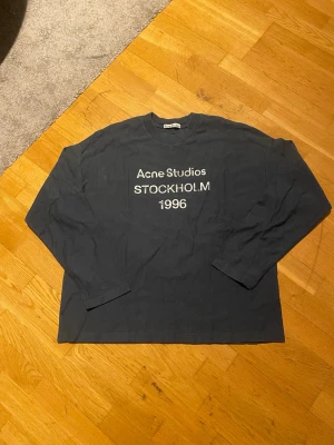 Acne Studios blå sleeve - Cool mörkblå långärmad topp från Acne Studios med tryck 'STOCKHOLM 1996' på bröstet. Klassisk rund halsringning och relaxed fit. Perfekt till jeans eller kjol för en enkel men snygg look.