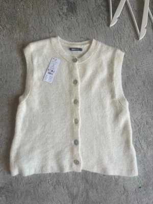 Vit stickad väst från Gina Tricot - Mysig vit stickad väst från Gina Tricot med rund halsringning och knappar framtill. Västen har en enkel och clean look som passar perfekt till lager-på-lager outfits. Tillverkad i mjukt material som känns skönt mot huden. Helt ny aldrig använd.
