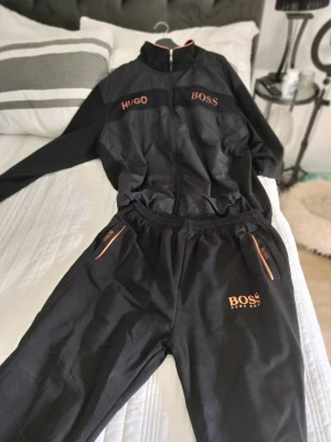 Svart Hugo Boss ziptröja med orange detaljer - Snygg svart ziptröja från Hugo Boss med orange logga och detaljer. Tröjan har hög krage, dragkedja framtill och matchande orangea dragkedjor på fickorna. Perfekt för en sportig och avslappnad stil. Köpt utlandet