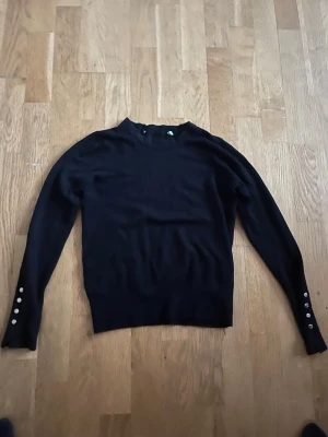 Svart stickad tröja med knappar - Snygg svart stickad tröja med rund hals och långa ärmar. Ärmsluten har dekorativa guldfärgade knappar som ger en extra detalj. Perfekt till jeans eller kjol för en stilren look.
