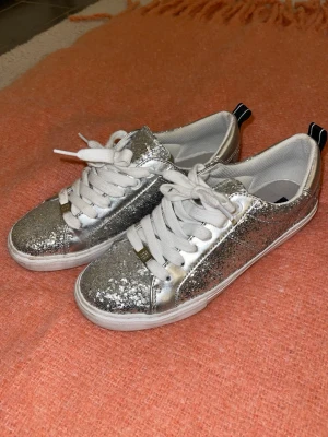 Silvriga glittriga sneakers - Säljer ett par riktigt snygga sneakers i silver med glitterdetaljer. Skorna är hela, inprencip oanvända, det ända är smittset på det ena sko snöret men det går att tvätta bort!