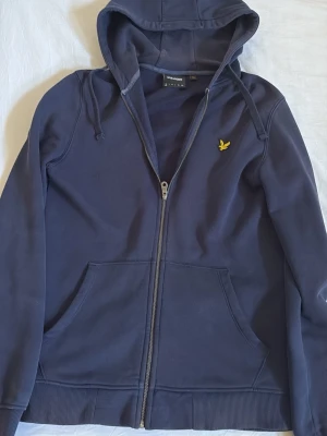 Marinblå hoodie från Lyle & Scott - Snygg marinblå hoodie från Lyle & Scott med dragkedja framtill, två stora fickor och klassisk gul logga på bröstet. Modellen har huva med dragsko och ribbade muddar. Perfekt för dig som gillar stilrena och bekväma plagg.