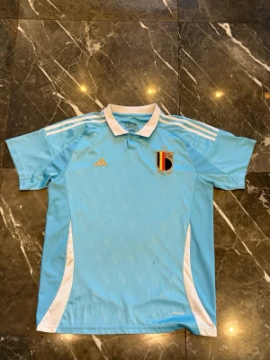 Belgien Adidas ljusblå matchtröja - Säljer en ljusblå matchtröja från Adidas med Belgien landslagets emblem på bröstet. Tröjan har vit krage, vita detaljer längs sidorna och axlarna samt guldigt Adidas-märke. Perfekt för fotbollsträning eller att bära till matchen.
