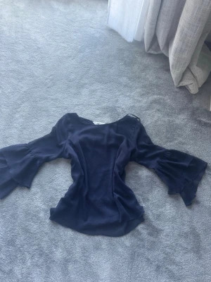 Mörkblå blus med volangärmar - Snygg mörkblå blus med vida volangärmar och rund halsringning. Tillverkad i ett lätt, flytande tyg som ger en elegant siluett. Perfekt för dig som vill ha en trendig och stilren look med extra detaljer på ärmarna.