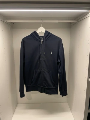 Mörkblå hoodie från Polo Ralph Lauren - Snygg mörkblå hoodie med dragkedja från Polo Ralph Lauren. Klassisk design med vit logga på bröstet, två fickor fram och justerbar huva med snören. Perfekt för en avslappnad och stilren look.