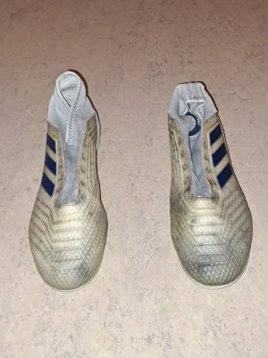 Adidas vita fotbollsskor med blå ränder - Säljer ett par vita fotbollsskor från Adidas med klassiska blå ränder på sidorna. Skorna har en strumpliknande design utan snörning och mönstrad ovandel för extra grepp. Yttersulan har fasta dobbar för spel på gräs. Perfekta för dig som vill ha stilrena och funktionella skor på planen. I bra skick. Säljs för 90 kr. Tveka inte att kontakta om du är intresserad eller har några frågor! Först till kvarn dom gäller!