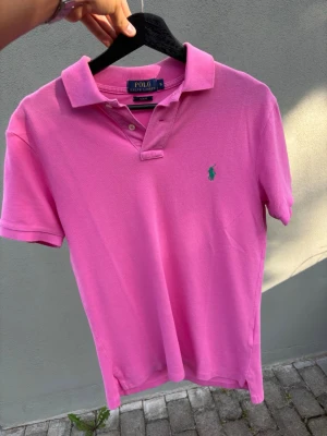 Rosa pikétröja från Polo Ralph Lauren - Säljer en rosa pikétröja från Polo Ralph Lauren i slim fit. Tröjan har klassisk krage, korta ärmar och den ikoniska gröna logon broderad på bröstet. Perfekt för en avslappnad men ändå stilren look.