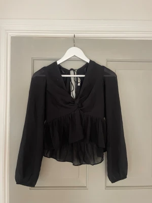 Svart tunn blus med knyt - Stilren svart blus i transparent material med v-ringning och knytband både fram och bak. Blusen har volang nedtill och långa ärmar med lätt puff. Man kan både klä upp och klä ner den!