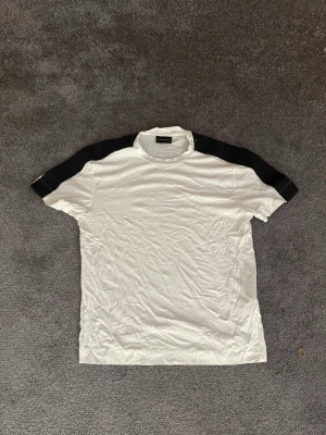 Armani T-shirt - Snygg vit t-shirt från Emporio Armani med svarta paneler över axlarna. Klassisk rund hals och korta ärmar. Tillverkad i mjuk bomull som känns skön mot huden. Perfekt för en clean och stilren look. Storlek S i väldigt fint skick. 