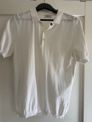 Vit stickad kortärmad polotopp - Säljer en vit stickad polotopp med korta ärmar och krage. Toppen har knappar framtill och är tillverkad i ett lätt, strukturerat material som ger en avslappnad men ändå stilren look. Perfekt för varmare dagar och enkel att matcha med olika outfits.