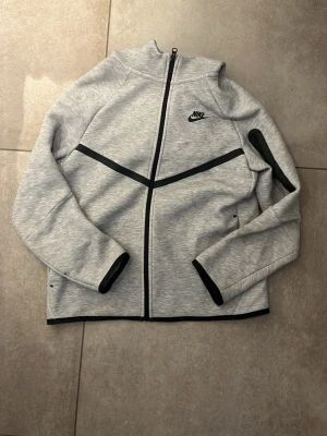 Nike tech fleece grå - Snygg grå hoodie från Nike med hel dragkedja framtill och svarta detaljer längs dragkedjan, ärmslut och bröstet. Klassisk Nike-logga på bröstet och huva. Perfekt för en sportig och avslappnad stil. Har blivit hål som har lagats men inget som är störande.