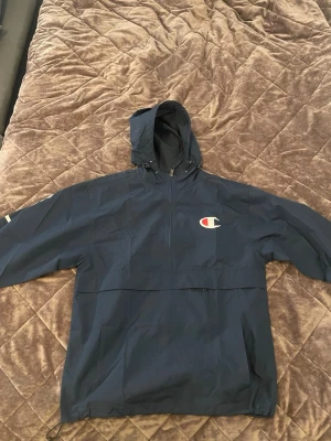Champion Windbreaker Anorak – Navy / Retro - Mörkblå Champion windbreaker/anorak med den klassiska stora Champion C-loggan på bröstet. Sportig retrodesign som passar perfekt till streetwear och vardags.  Endast använd 1 gång – i nyskick!  • Nypris: 599 kr • Champion • Mörkblå / Navy • Windbreaker / Anorak • Half-zip • Huva med dragsko • Stor Champion C-logo • Frontficka • Retro / streetwear-look  Lätt och smidig jacka som passar perfekt under vår, sommar och höst.  Skickas snabbt efter köp 📦