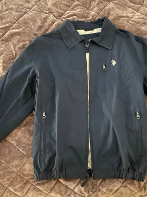 U.S. Polo Assn Retro Jacka – Mörkblå - U.S. Polo Assn jacka i mörkblå färg. Stilren retro-modell som inte längre säljs i butik, vilket gör den lite mer unik.  Jackan har klassisk krage, dragkedja framtill, tre fickor med dragkedja samt den broderade U.S. Polo Assn-loggan på bröstet.  Nypris: 1 299 kr Modell: Retro / utgången modell Färg: Mörkblå  Snygg både till vardags och som en tunnare vår-/höstjacka. Bra skick.  Skickas snabbt efter köp 📦