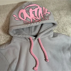 Syna world hoodie - Helt ny oanvänd SYNA WORLD hoodie i storlek S. Säljer den för att den är för liten för mig. aldrig använd, aldrig tvättad. —————————Helt stilren. Snygg grå hoodie med huva och rosa snören. På huvan finns ett stort rosa graffiti-inspirerat tryck som ger en streetwear vibe. Hoodien är gjord i mjukt material och har en avslappnad passform. Perfekt för dig som gillar unika detaljer och bekväma plagg.