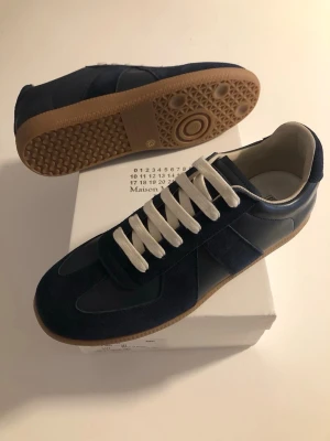 Maison Margiela - Snygga mörkblå sneakers med vita snören och beige gummisula. Skorna har en klassisk låg siluett och är tillverkade i en mix av mocka och läder. Perfekta för dig som gillar stilrena och tidlösa sneakers.