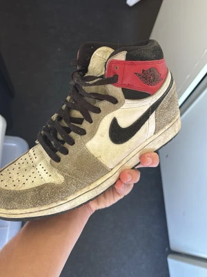 Nike Air Jordan 1 Retro High OG - Säljer ett par Nike Air Jordan 1 Retro High OG sneakers i vitt, svart och rött. Skorna har svart swoosh, svarta skosnören och röd detalj vid ankeln med klassisk Air Jordan-logga. Ovandelen är i mocka och läder för en ikonisk look.