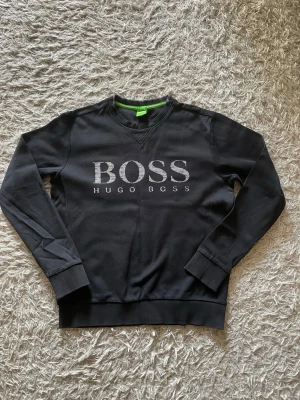 Svart sweatshirt från Hugo Boss - Snygg svart sweatshirt från Hugo Boss med stort BOSS-tryck i vitt på bröstet. Tröjan har rund halsringning, ribbade muddar vid ärmslut och nederkant samt en grön detalj i nacken. Perfekt för en stilren och avslappnad look.