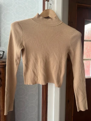 Beige ribbad stickad polotröja - Trendig beige stickad tröja med ribbad struktur och hög krage. Modellen är croppad och har långa ärmar. Perfekt att matcha med höga jeans eller kjol för en clean och stilren look.