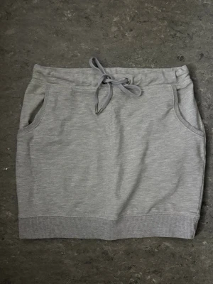 Grå kortkjol med snörning - Mjuk och bekväm grå kortkjol i sweatshirtmaterial med snörning i midjan och två fickory fram. Kjolen har ribbad kant nedtill och en avslappnad passform som passar perfekt till vardagslooken.